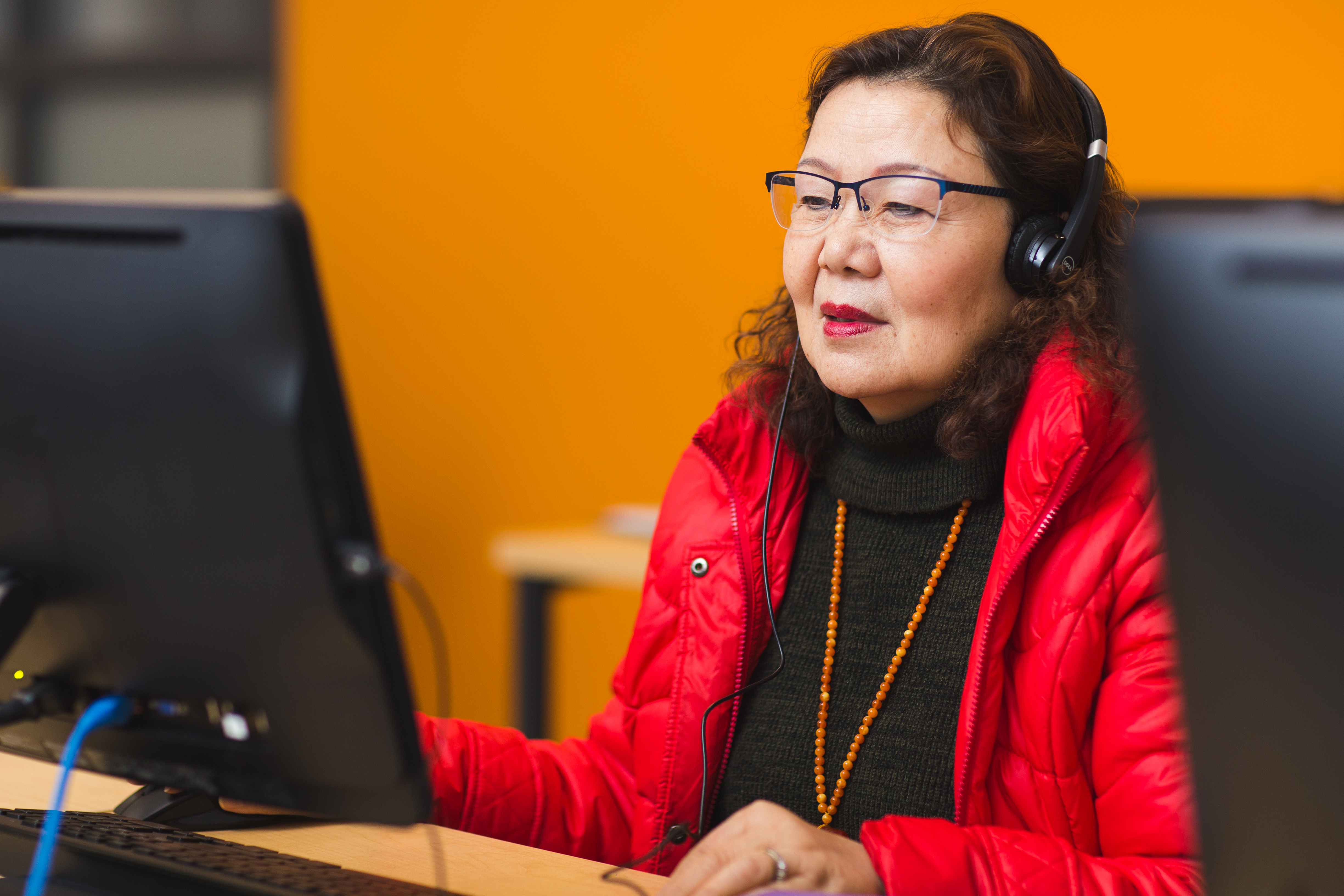 A woman in a black shirt, red coat, and glasses performs language work on a desktop computer.