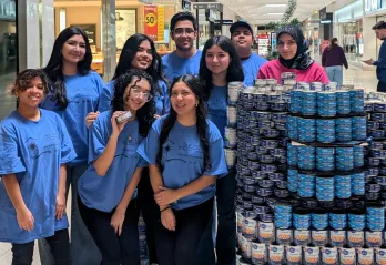 A group of nine young volunteers wearing matching blue t-shirts with "The Inn of The Good Shepherd" logo stand together in a shopping mall, smiling at the camera. They are positioned next to a large, neatly stacked display of canned food, primarily tuna and chickpeas. One volunteer in the front row holds up a can. In the background, shoppers walk through the mall, and a sign for the YMCA Canned Food Drive is visible.