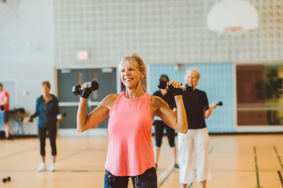 An older woman in a pink tank top smiles and lifts weights as part of a group fitness class.