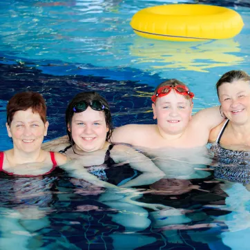 A family of four (two women, a girl and boy) smile and hug in a YMCA pool.