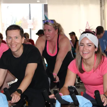 A group of people in pink and black shirts cycle on stationery bikes at a SWEAT for Strong Kids event.