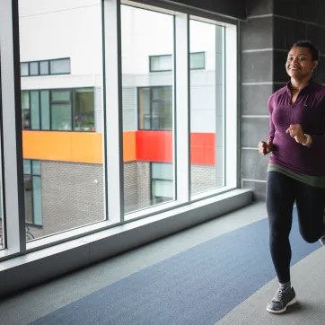 A woman in a purple shirt and black leggings jogs on an indoor walking track.