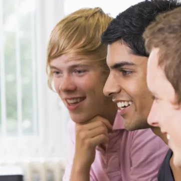 Three adolescents laugh as they play a game online in front of computer monitors.